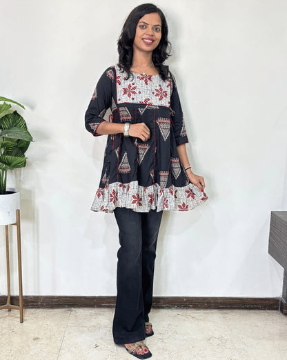 Black Women's Ajrakh Vegetable Dyed Plant based Cotton Short Kurti with three quarter sleeves . Grey & Black base & geometric print on Sleeves & front , Standing in front of white wall. 