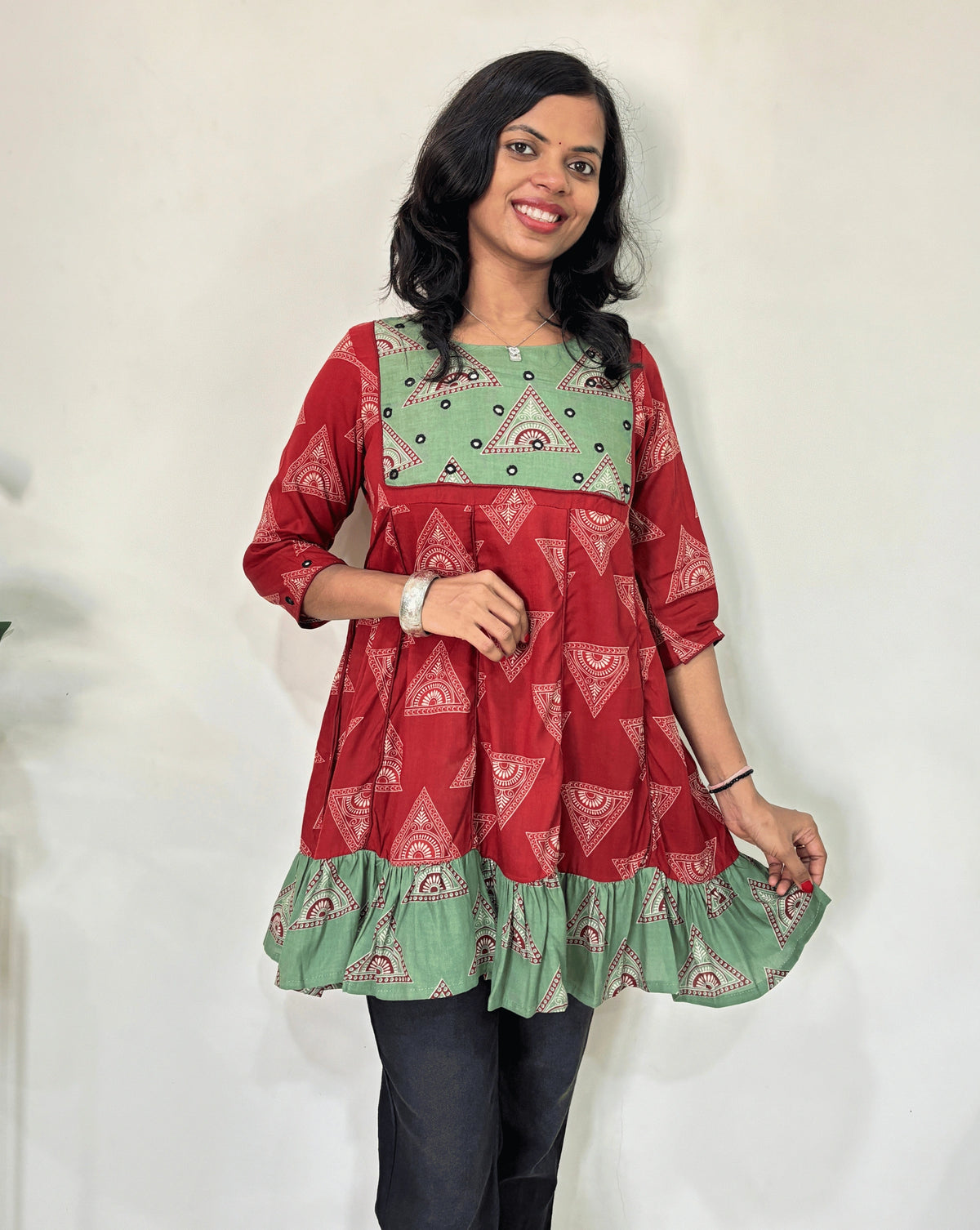 Front veiw of Red Ajrakh Plant based cotton Short kurti featured with Red and Green geometric print & aline frill patterned dress,  standing against a white wall.