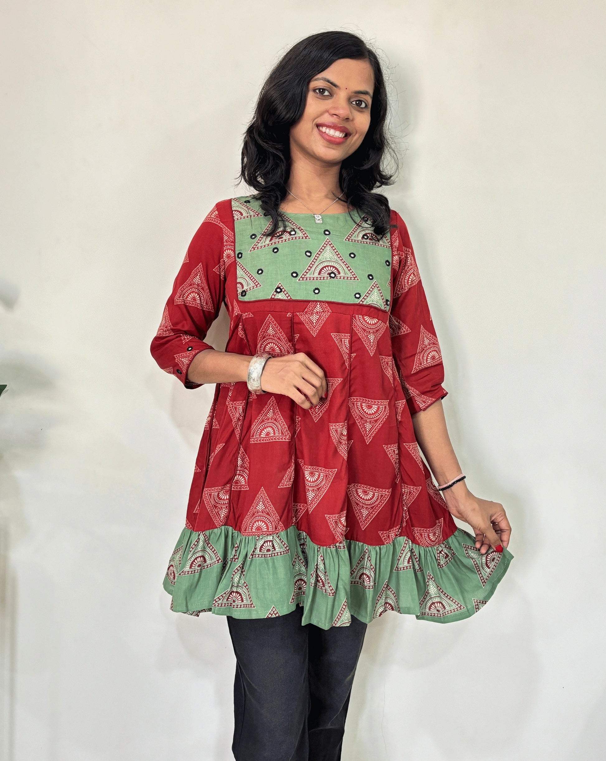 Front veiw of Red Ajrakh Plant based cotton Short kurti featured with Red and Green geometric print & aline frill patterned dress,  standing against a white wall.