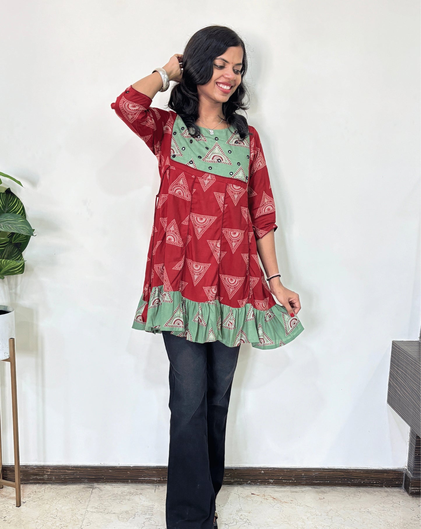 Red Women's Ajrakh Vegetable Dyed Plant based Cotton Short Kurti with three quarter sleeves . Green & Red base & geometric print on Sleeves & front , Standing in front of white wall. 