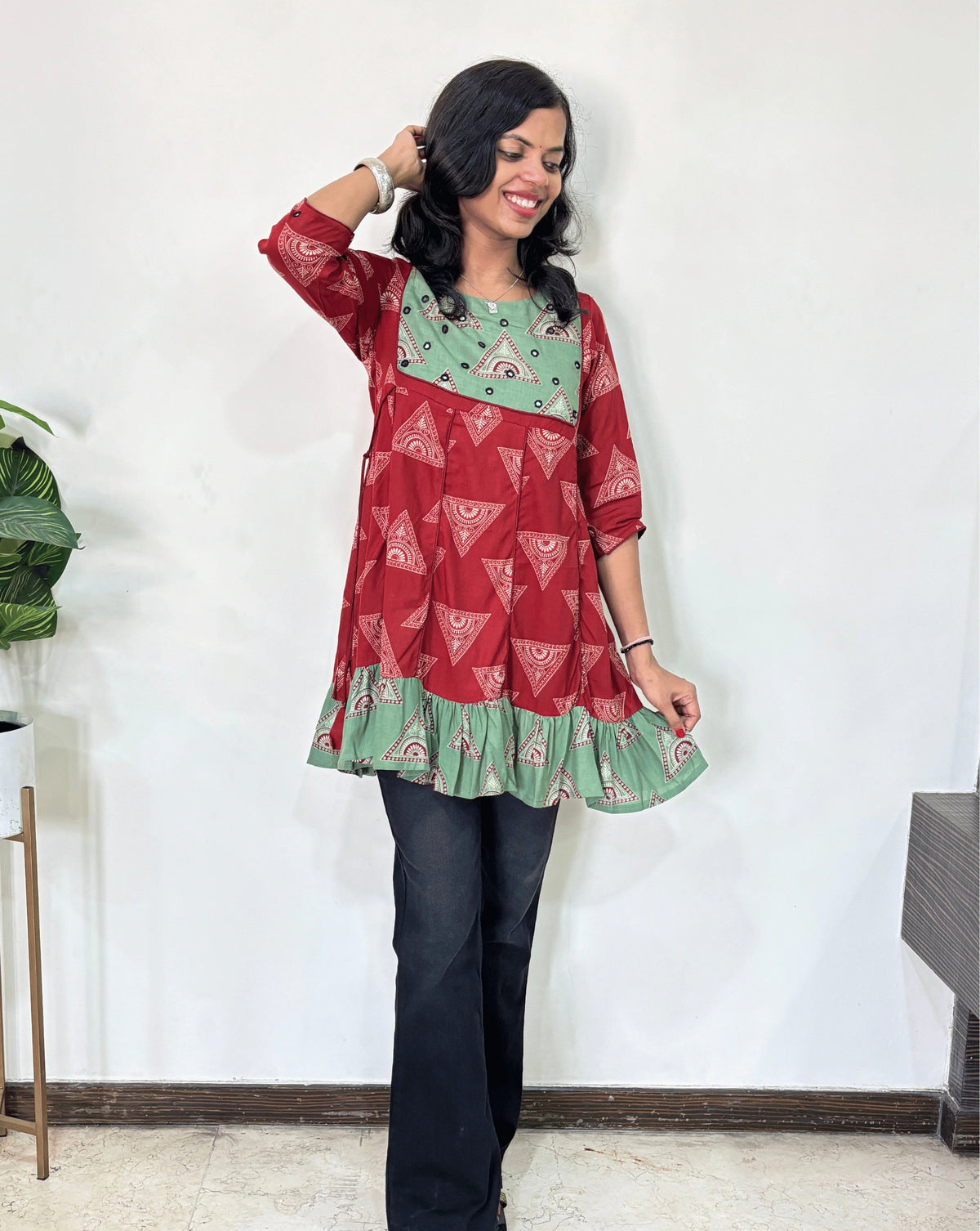 Red Women's Ajrakh Vegetable Dyed Plant based Cotton Short Kurti with three quarter sleeves . Green & Red base & geometric print on Sleeves & front , Standing in front of white wall. 