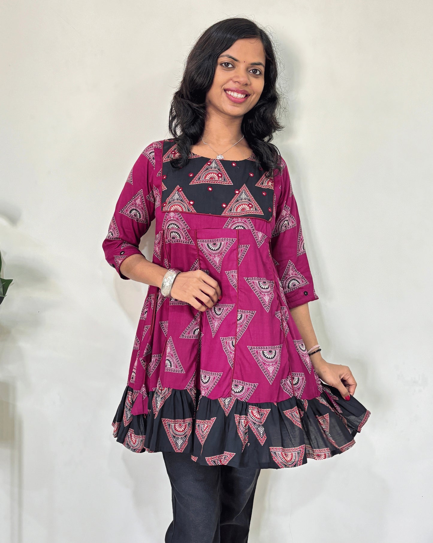 Close-up of Magenta Ajrakh plant based cotton Short kurti with geometric patterns, a Black brush print yoke & bottom, Red panel strip with lace on side, Hand-mirror work on round neck paired with dark pants against a plain light background.