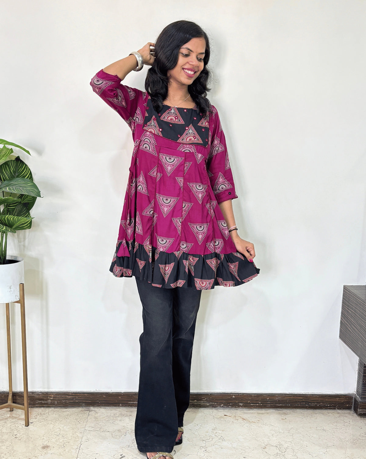 Front veiw of Magenta Ajrakh Plant based cotton Short kurti featured with pink and black geometric print & aline frill patterned dress,  standing against a white wall.