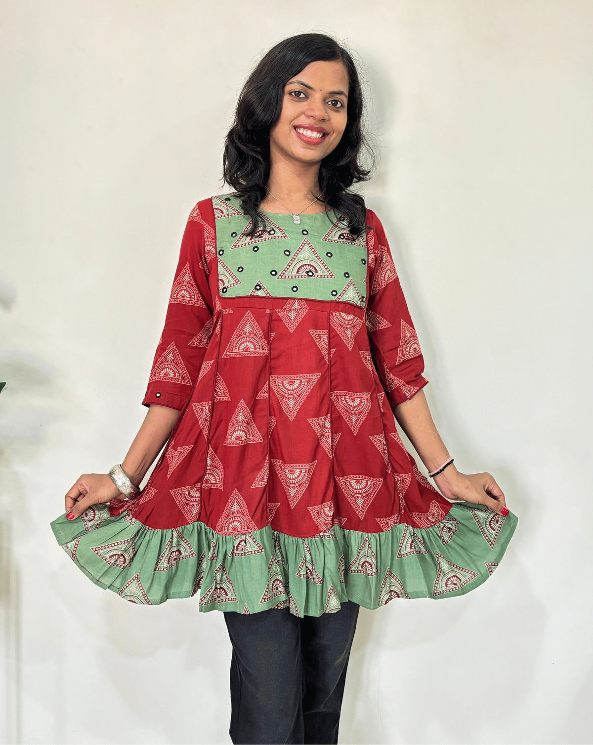 Close-up of Red Ajrakh plant based cotton Short kurti with geometric patterns, a Green brush print yoke & bottom, Red panel strip with lace on side, Hand-mirror work on round neck paired with dark pants against a plain light background.