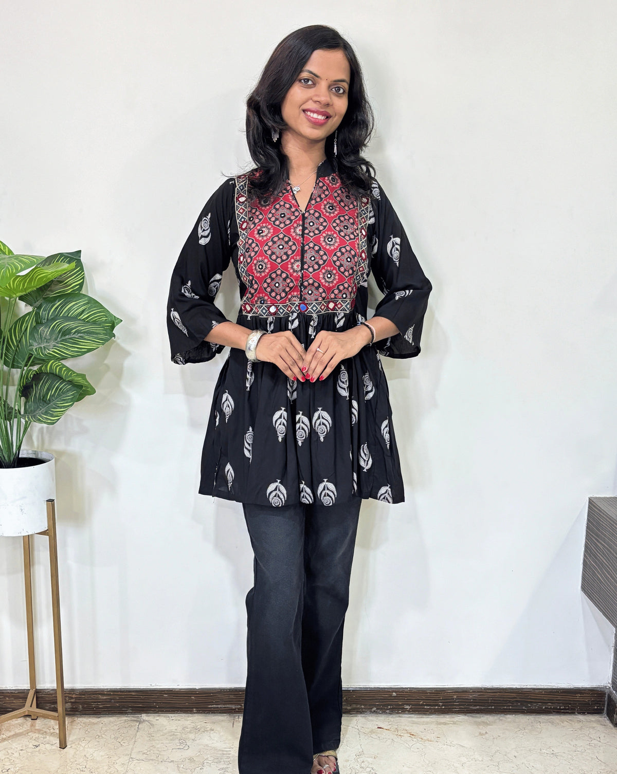 Front view of Black Ajrakh plant based cotton Short Kurti  with red base & geometric print yoke, leaf print on black , black pants in a room with a plant and white wall. 