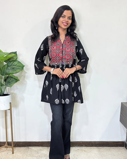 Front view of Black Ajrakh plant based cotton Short Kurti  with red base & geometric print yoke, leaf print on black , black pants in a room with a plant and white wall. 
