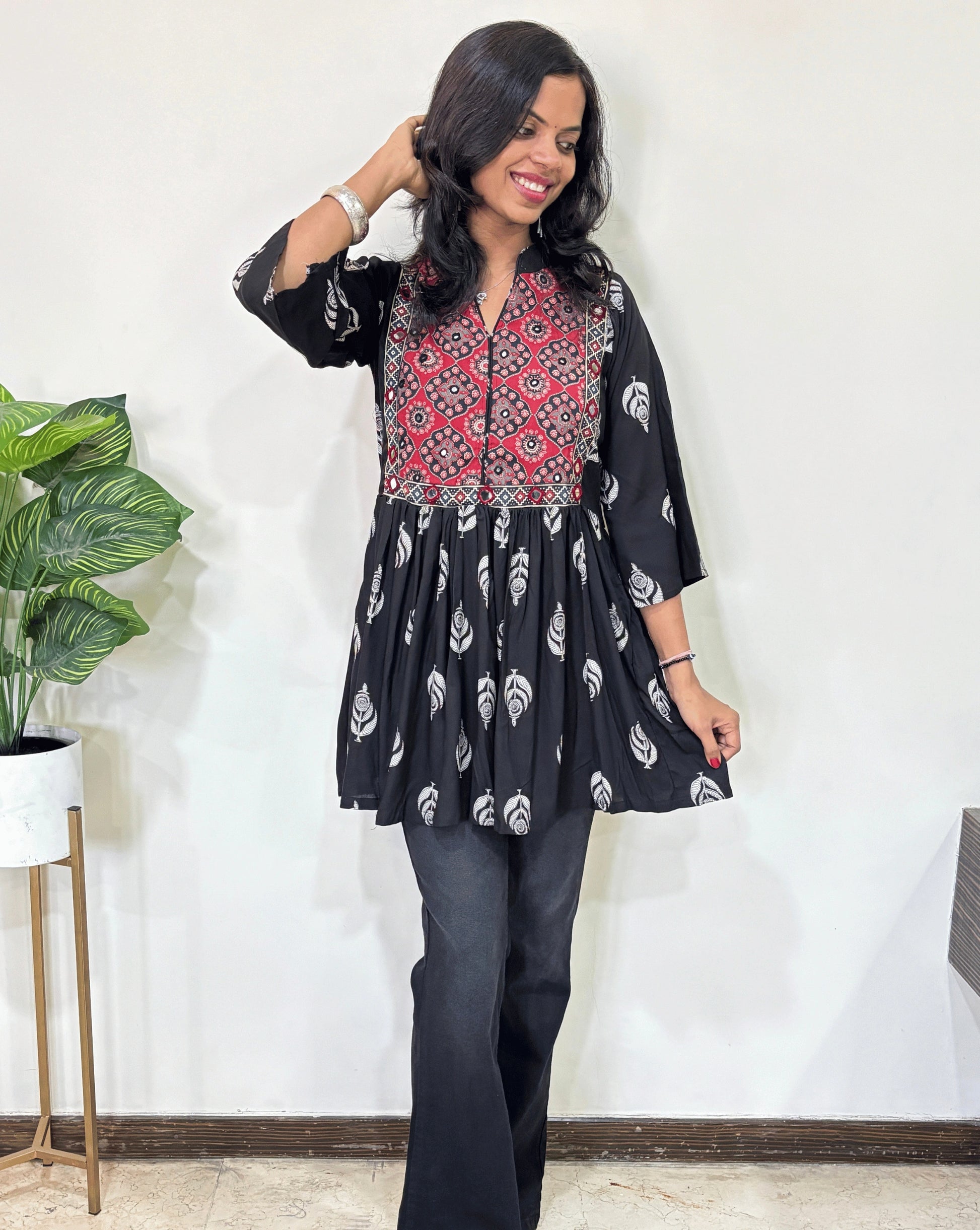 Black women's Ajrakh vegetabled dyed plant based Cotton Short Kurti featured with three quarter Loose sleeves,  Black and red leaf print  patterned ,standing indoors with a plant in the background.