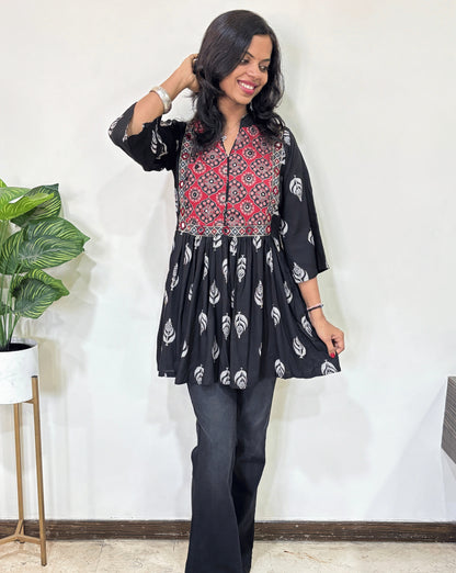 Black women's Ajrakh vegetabled dyed plant based Cotton Short Kurti featured with three quarter Loose sleeves,  Black and red leaf print  patterned ,standing indoors with a plant in the background.
