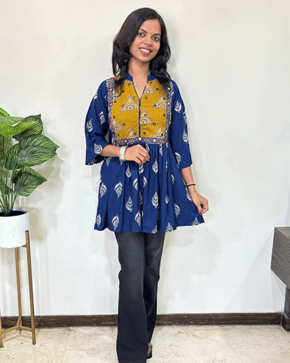 Front view of  blue Ajrakh plant based cotton Short Kurti with yellow base & geometric print yoke, leaf print on black , black pants in a room with a plant and white wall. 