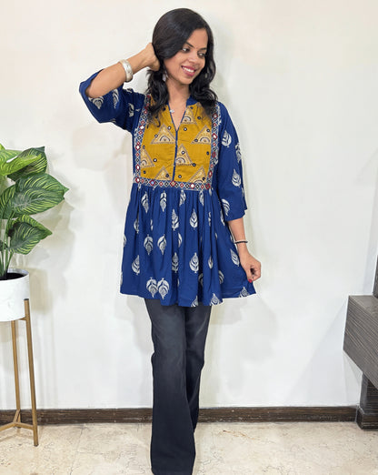 Navy-blue women's Ajrakh vegetabled dyed plant based Cotton Short Kurti featured with three quarter Loose sleeves,  Blue and mustard yellow ,leaf print  patterned ,standing indoors with a plant in the background.