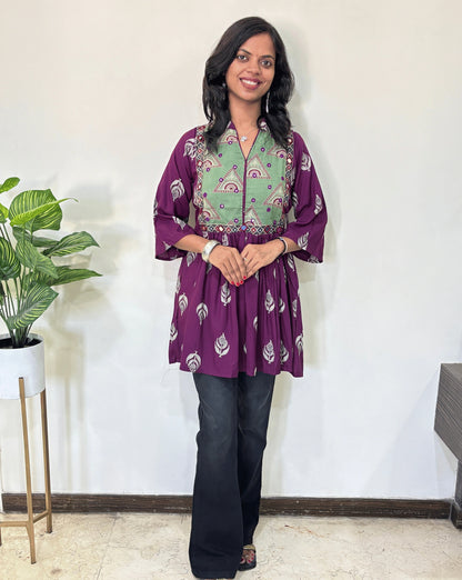 Front view of  Purple Ajrakh plant based cotton Short Kurti with Green base & geometric print yoke, leaf print on black , black pants in a room with a plant and white wall. 