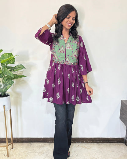 Purple women's Ajrakh vegetabled dyed plant based Cotton Short Kurti featured with three quarter Loose sleeves,  purple and green leaf print  patterned ,standing indoors with a plant in the background.