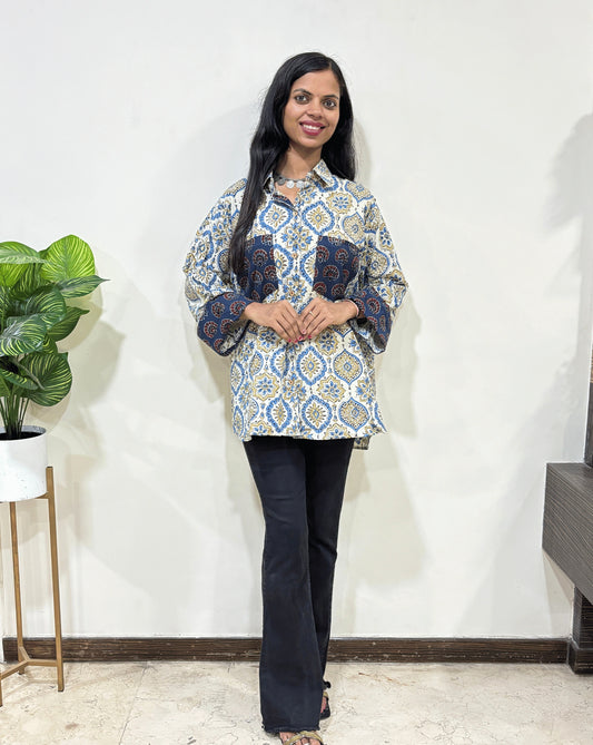 Front view of White Women's Ajrakh vegetable dyed plant based cotton Shirt with a Blue & yellow badam-buta print , 2 pocket in front Paired with black pants standing indoors with a white wall and plant in the background.