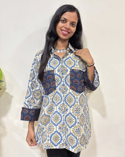 Close-up of White Women's Ajrakh vegetable dyed plant based cotton Shirt with a white & blue badam-buta print, 2 pocket in front, collar neck,  Paired with black pants standing indoors with a white wall and plant in the background.