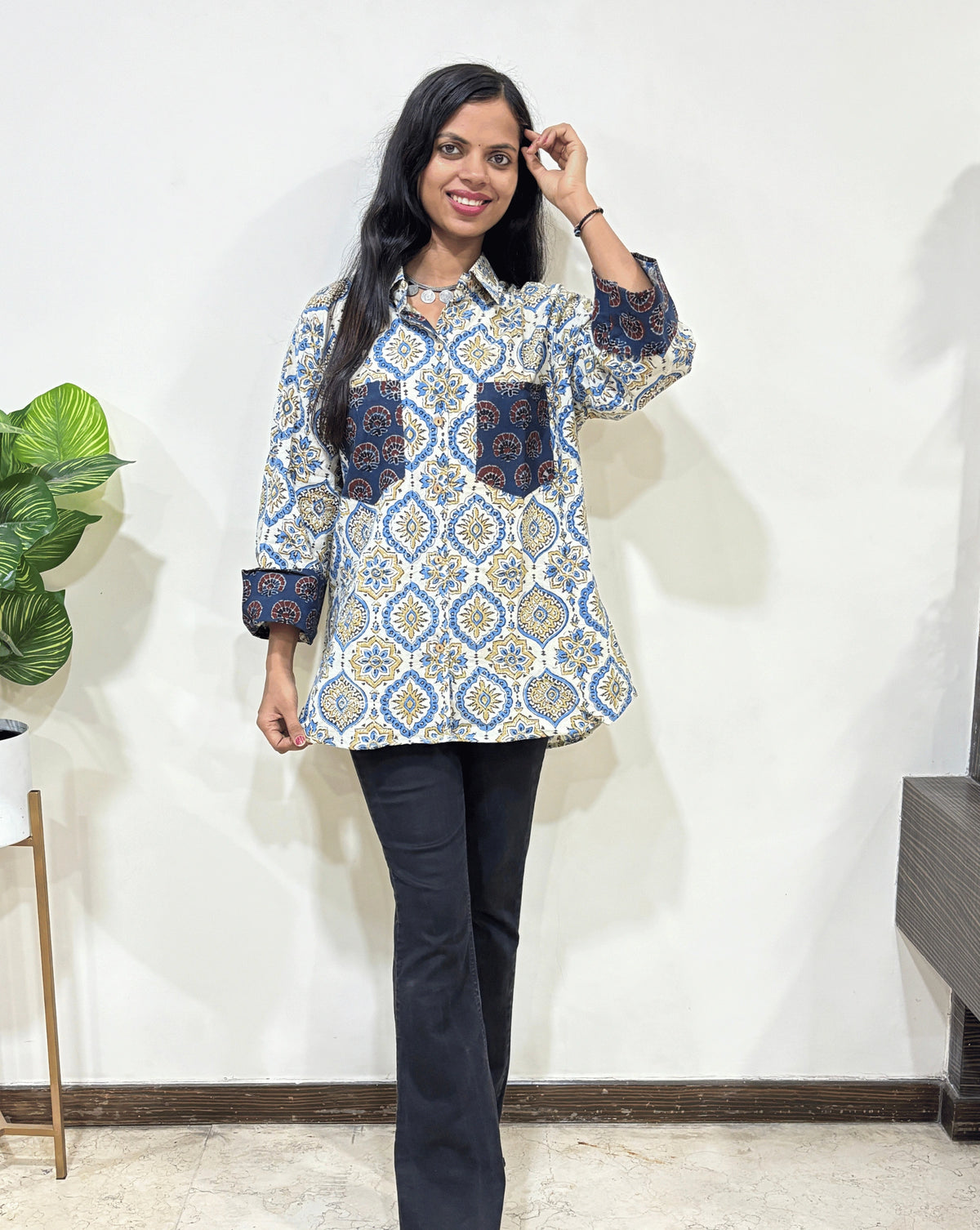 White Women's Ajrakh vegetable dyed plant based cotton Shirt with a blue & yellow badam-buta print , Paired with black pants standing indoors with a white wall and plant in the background.
