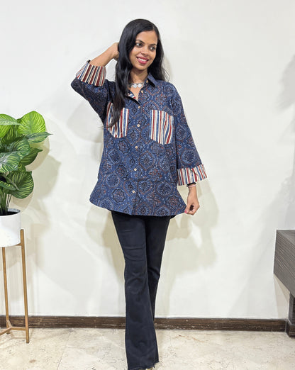 Blue Women's Ajrakh vegetable dyed plant based cotton Shirt with Geometric print , Paired with black pants standing indoors with a white wall and plant in the background.