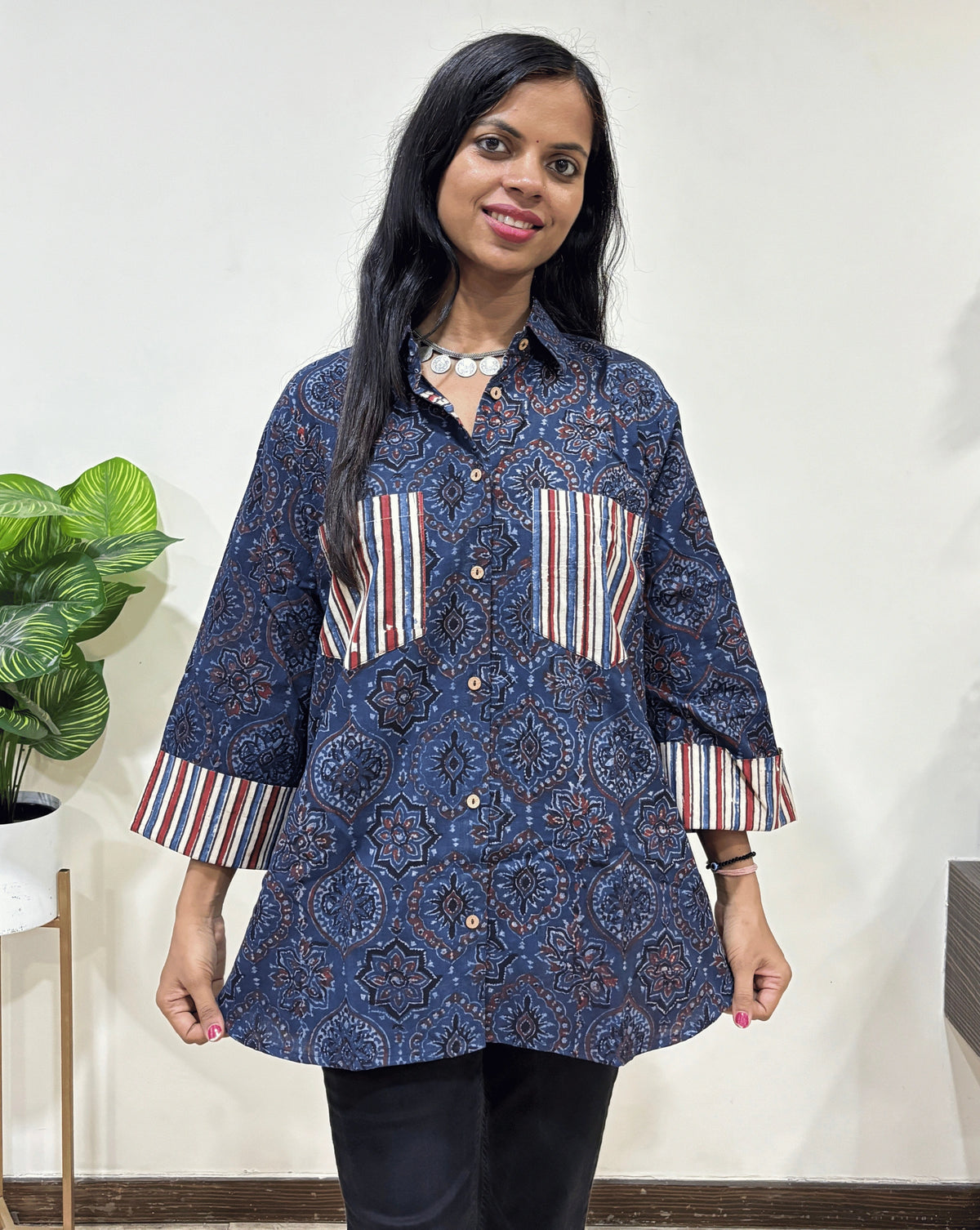 Front view of Blue Women's Ajrakh vegetable dyed plant based cotton Shirt with a Blue & yellow badam-buta print , 2 pocket in front Paired with black pants standing indoors with a white wall and plant in the background.
