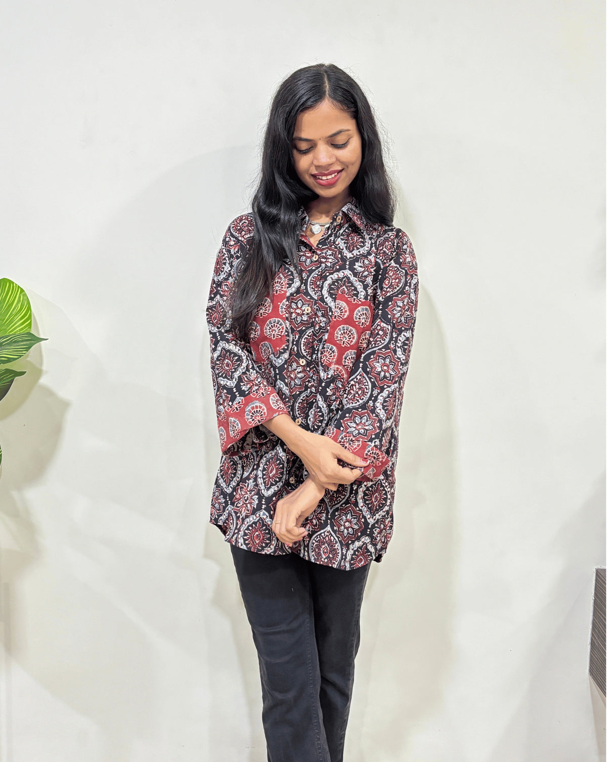 Close-up of Black Women's Ajrakh vegetable dyed plant based cotton Shirt with a Black & Red badam-buta print, 2 pocket in front, collar neck,  Paired with black pants standing indoors with a white wall and plant in the background.
