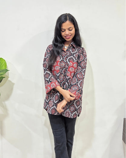 Close-up of Black Women's Ajrakh vegetable dyed plant based cotton Shirt with a Black & Red badam-buta print, 2 pocket in front, collar neck,  Paired with black pants standing indoors with a white wall and plant in the background.