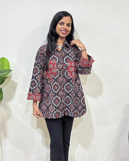 Front view of Black Women's Ajrakh vegetable dyed plant based cotton Shirt with a red & beige badam-buta print , 2 pocket in front Paired with black pants standing indoors with a white wall and plant in the background.