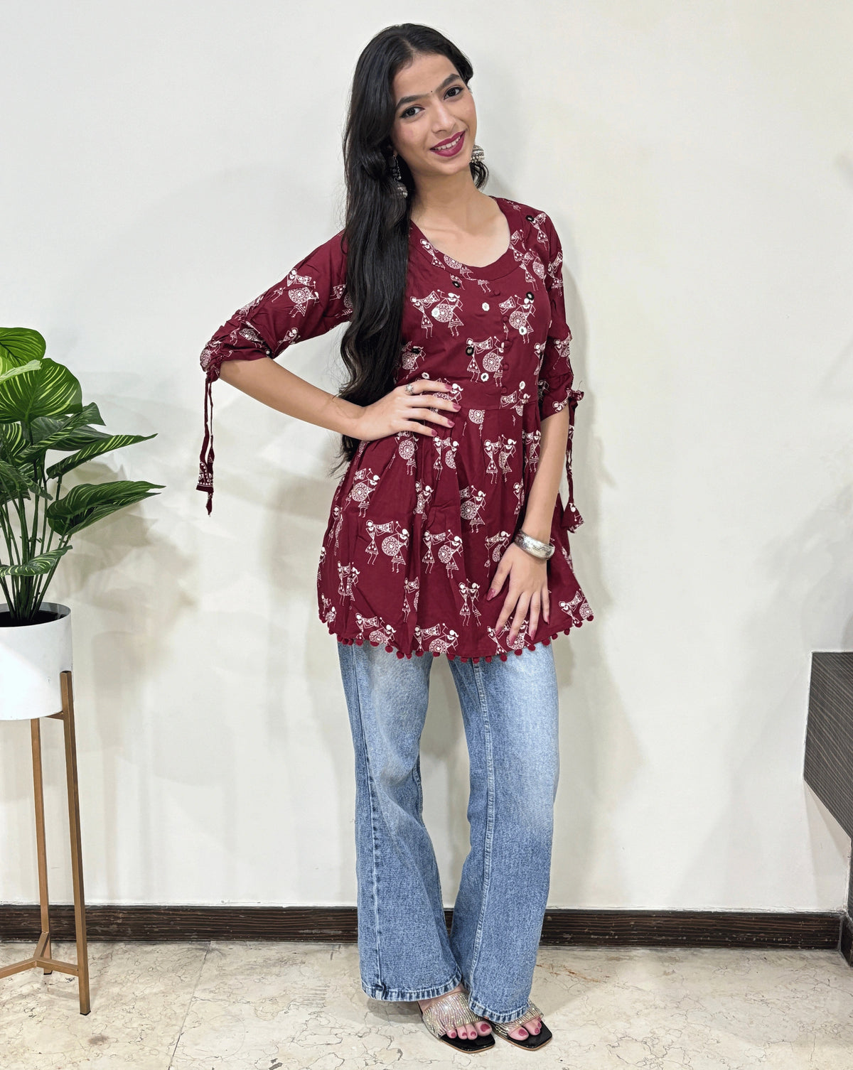 Front view of Red Woman's Ajrakh Vegetable dyed plant based cotton Short Kurti with beautiful Warli print & frock style pattern, lace tasels on sleeves, paired with light blue jeans, green leaf post on right side & Standing in front of white wall. 