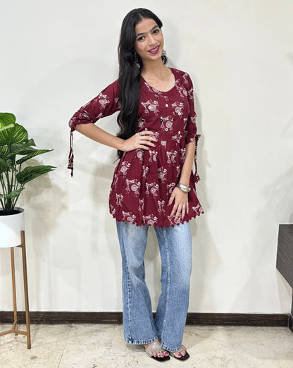 Front view of Red Woman's Ajrakh Vegetable dyed plant based cotton Short Kurti with beautiful Warli print & frock style pattern, lace tasels on sleeves, paired with light blue jeans, green leaf post on right side & Standing in front of white wall. 
