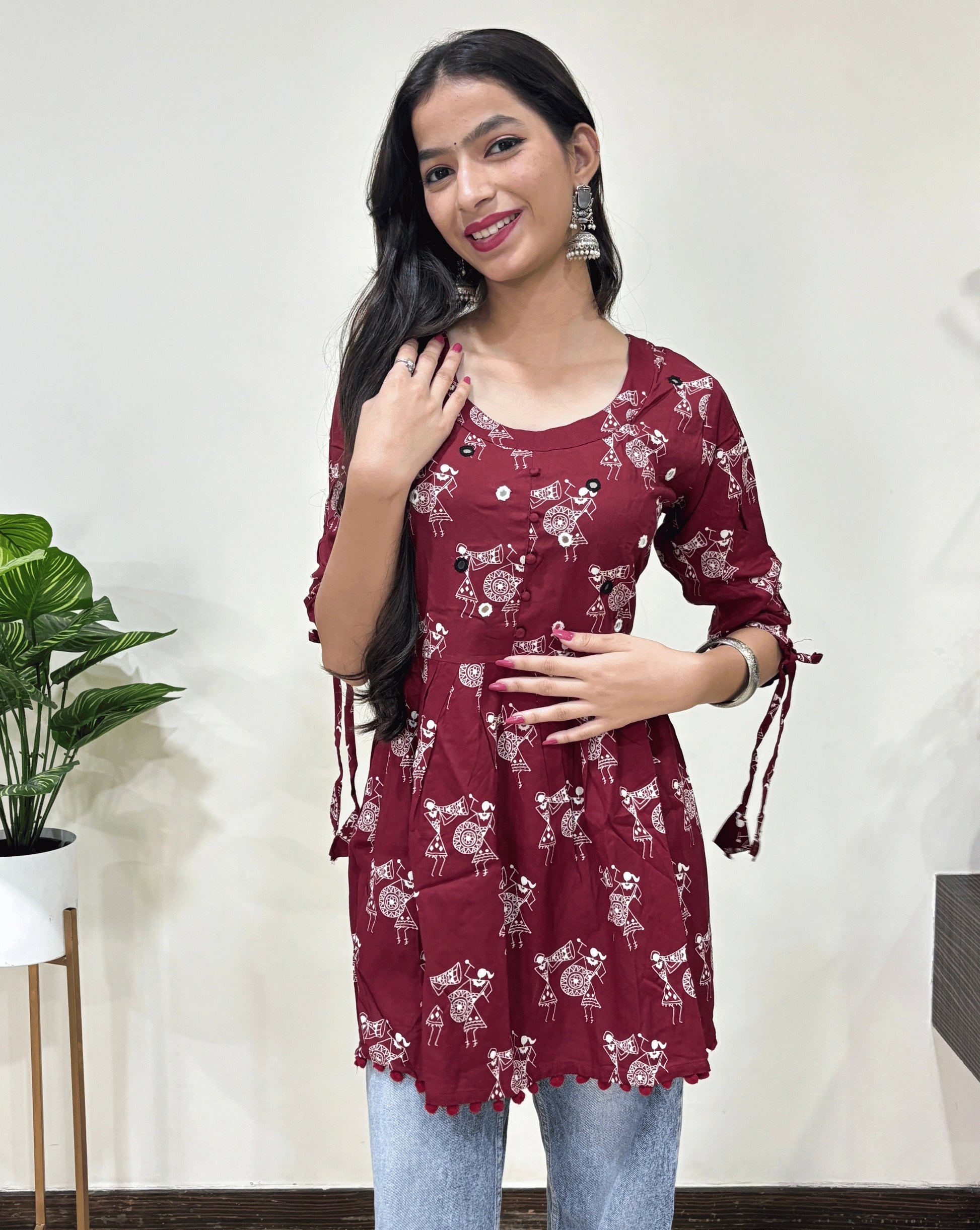 Close-up of Red Woman's Ajrakh Vegetable dyed plant based cotton Short Kurti with beautiful Warli print & frock style pattern, lace tasals on sleeves, small red potlis on front & bottom, round neck with hand-mirror work, paired with light blue jeans, green leaf post on right side & Standing in front of white wall. 