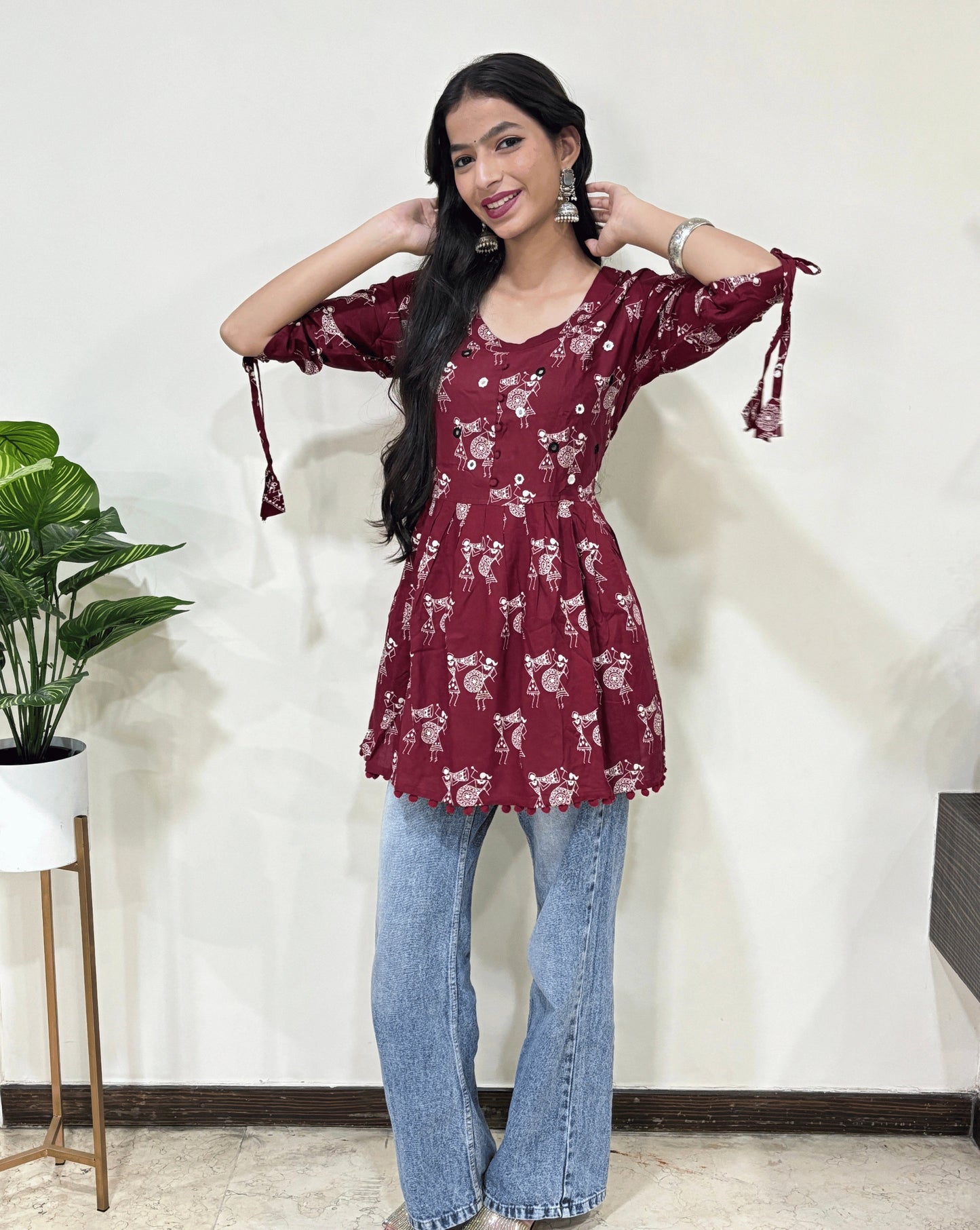 Red Woman's Ajrakh Vegetable dyed plant based cotton Short Kurti with beautiful Warli print & frock style pattern, paired with light blue jeans, green leaf post on right side & Standing in front of white wall. 