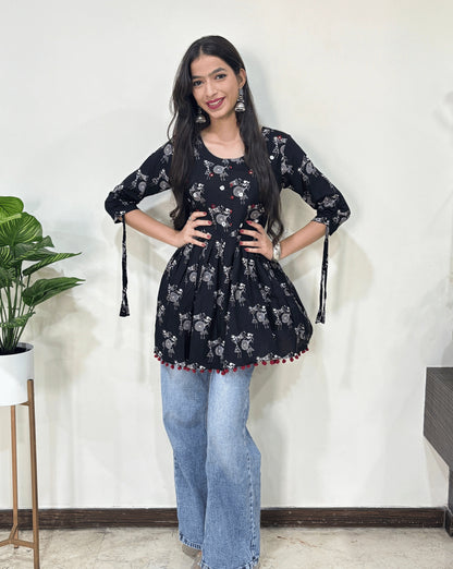 Front veiw of Black Woman's Ajrakh Vegetable dyed plant based cotton Short Kurti with beautiful Warli print & frock style pattern, lace tasals on sleeves, paired with light blue jeans, green leaf post on right side & Standing in front of white wall. 