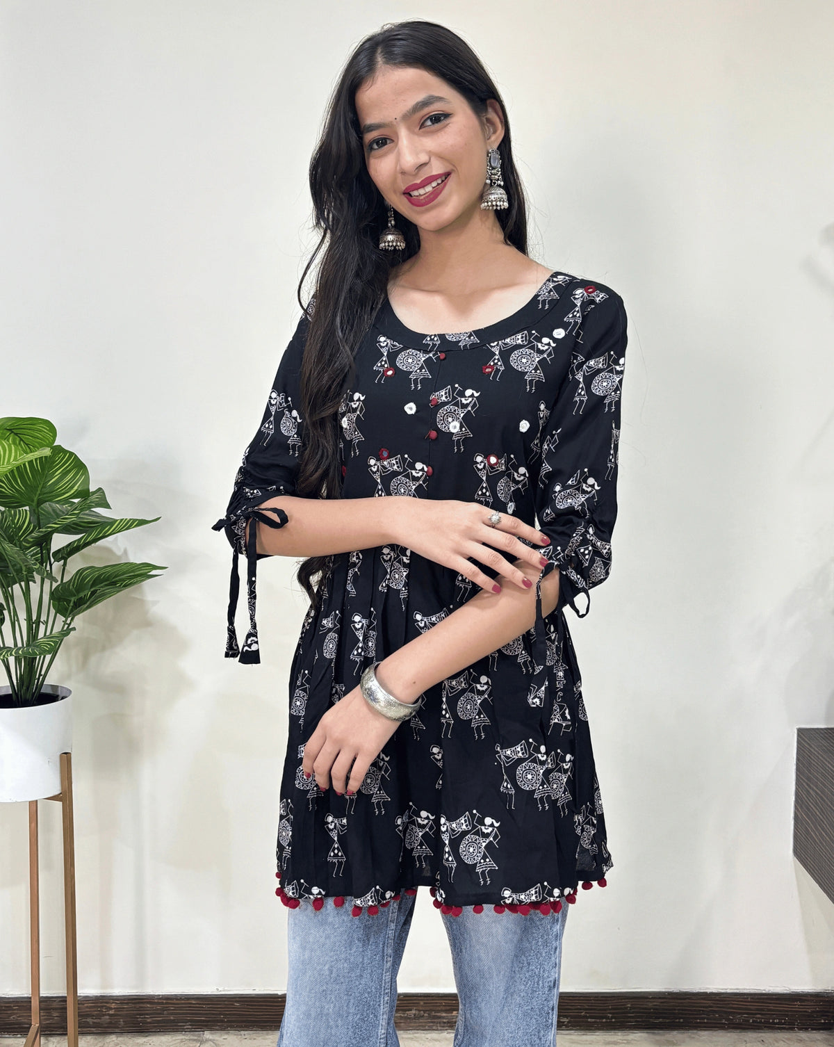 Close-up of Black Woman's Ajrakh Vegetable dyed plant based cotton Short Kurti with beautiful Warli print & frock style pattern, lace tasals on sleeves, small red potlis on front & bottom, round neck with hand-mirror work, paired with light blue jeans, green leaf post on right side & Standing in front of white wall. 