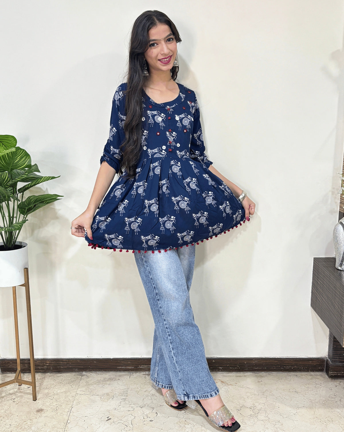 Blue Woman's Ajrakh Vegetable dyed plant based cotton Short Kurti with beautiful Warli print & frock style pattern, paired with light blue jeans, green leaf post on right side & Standing in front of white wall. 