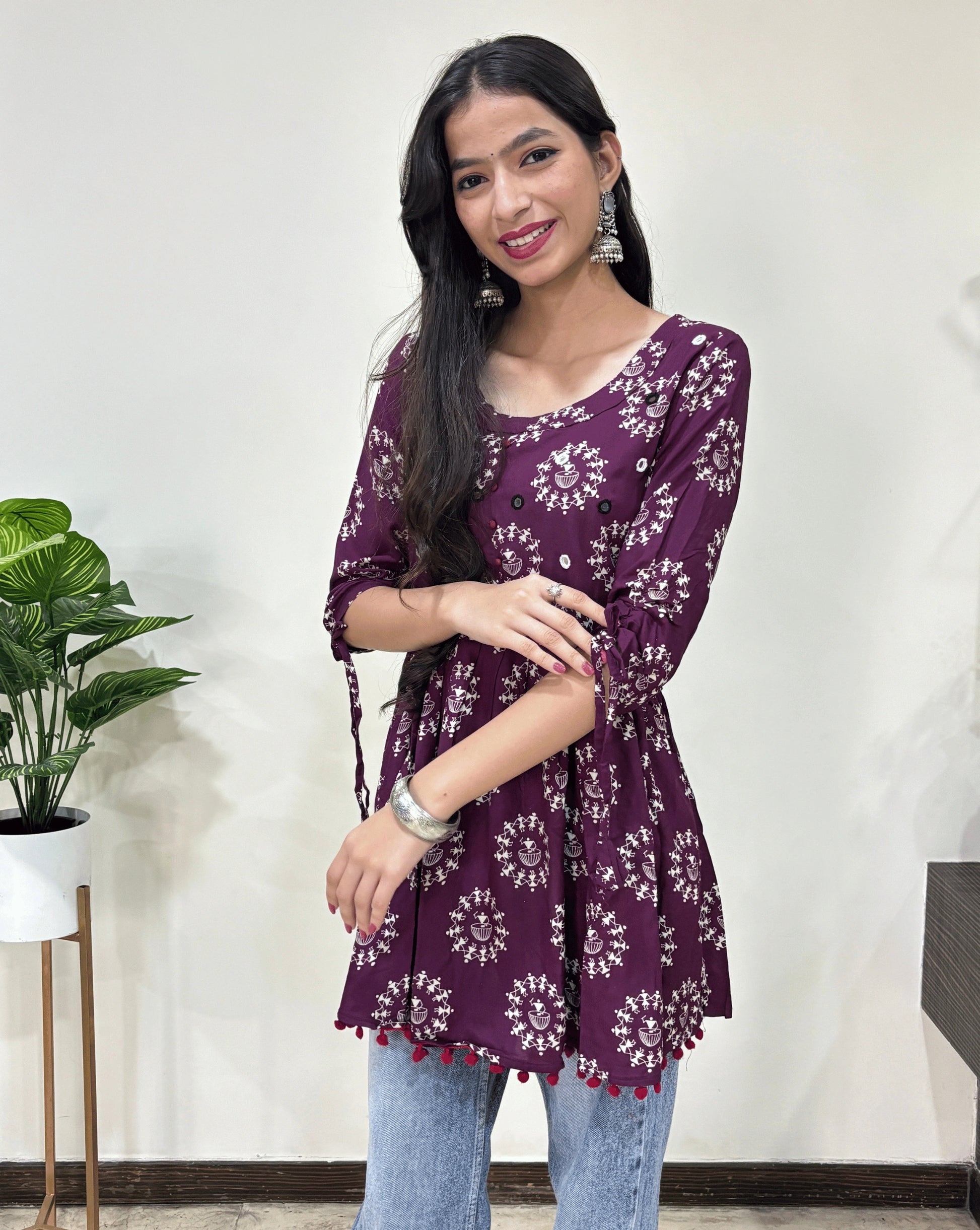 Front view of Purple Woman's Ajrakh Vegetable dyed plant based cotton Short Kurti with beautiful geometric Warli print, frock style pattern, Round neck with Hand-mirror work,  paired with light blue jeans, green leaf post on right side & Standing in front of white wall. 