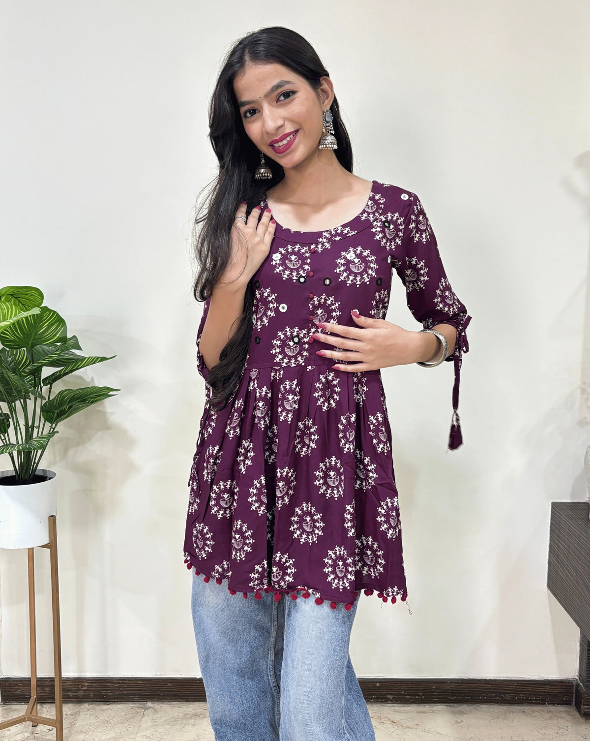 Close-up of Purple Woman's Ajrakh Vegetable dyed plant based cotton Short Kurti with beautiful geometric Warli print, frock style pattern, Round neck with Hand-mirror work, lace on sleeves,   paired with light blue jeans, green leaf post on right side & Standing in front of white wall. 