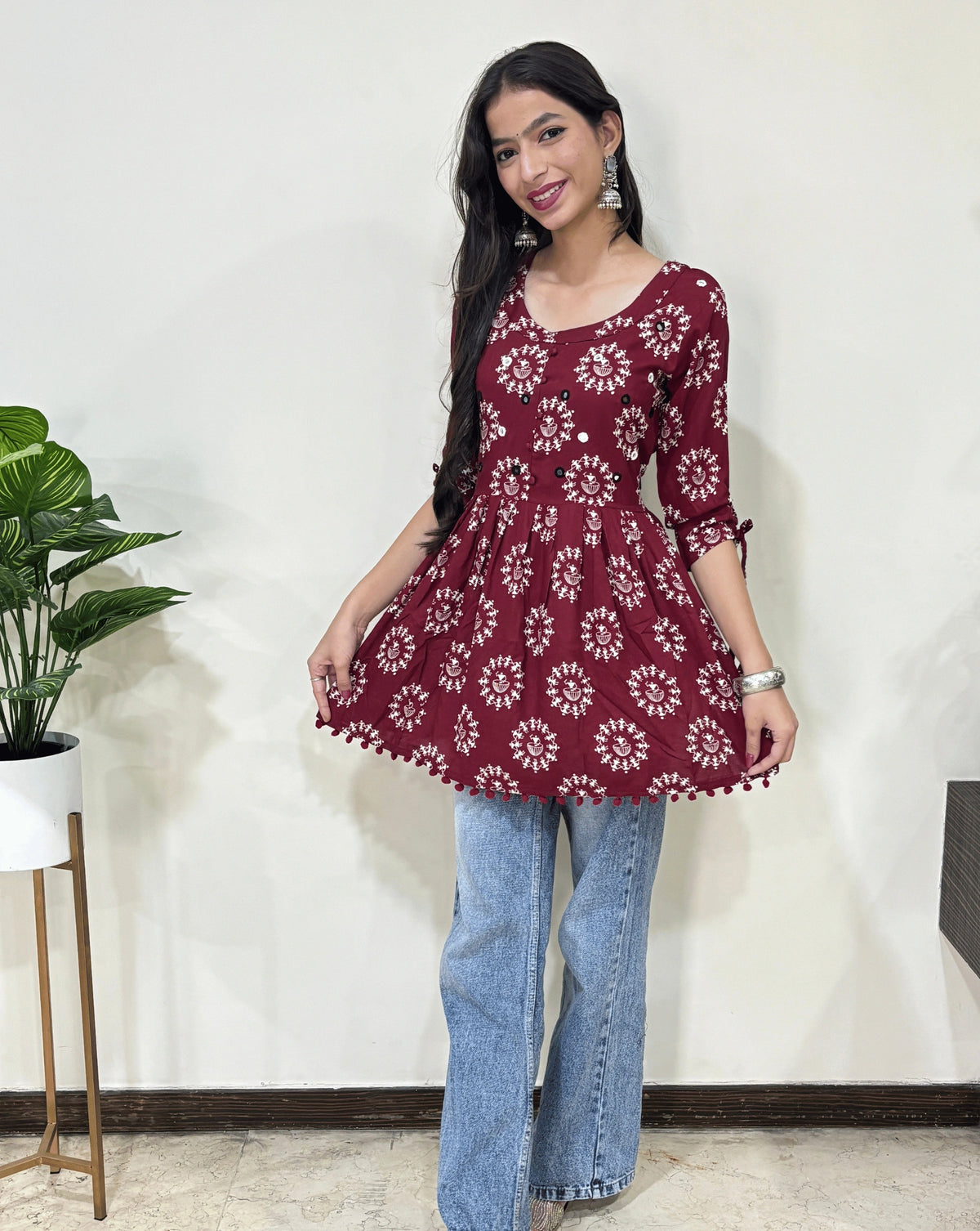 Front view of Red Woman's Ajrakh Vegetable dyed plant based cotton Short Kurti with beautiful geometric Warli print, frock style pattern, Round neck with Hand-mirror work,  paired with light blue jeans, green leaf post on right side & Standing in front of white wall. 