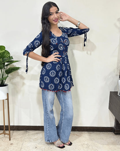 Blue Woman's Ajrakh Vegetable dyed plant based cotton Short Kurti with beautiful geometric Warli print, frock style pattern, paired with light blue jeans, green leaf post on right side & Standing in front of white wall. 