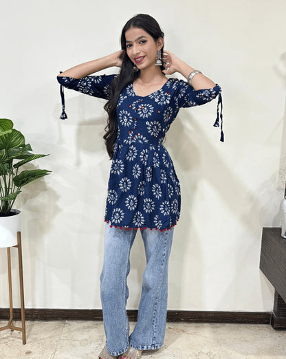Front view of Blue Woman's Ajrakh Vegetable dyed plant based cotton Short Kurti with beautiful geometric Warli print, frock style pattern, Round neck with Hand-mirror work,  paired with light blue jeans, green leaf post on right side & Standing in front of white wall. 