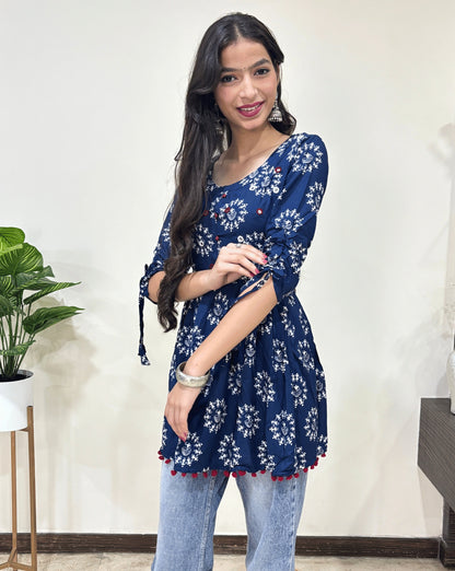 Close-up of Blue Woman's Ajrakh Vegetable dyed plant based cotton Short Kurti with beautiful geometric Warli print, frock style pattern, Round neck with Hand-mirror work, lace on sleeves,   paired with light blue jeans, green leaf post on right side & Standing in front of white wall. 