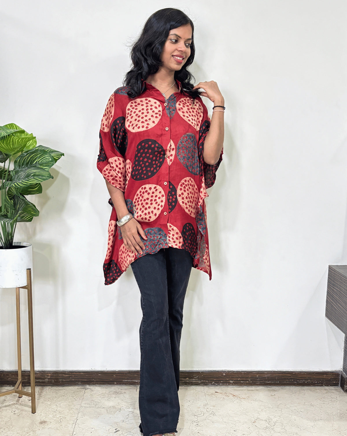 Front view of Red Women's Ajrakh pure Mashru Silk Short Kaftan Shirt Featured with Collar neck & Elbow sleeves with a bold abstract of pink, black, and red geometric circles print, paired with dark pants, standing in front of a white wall.