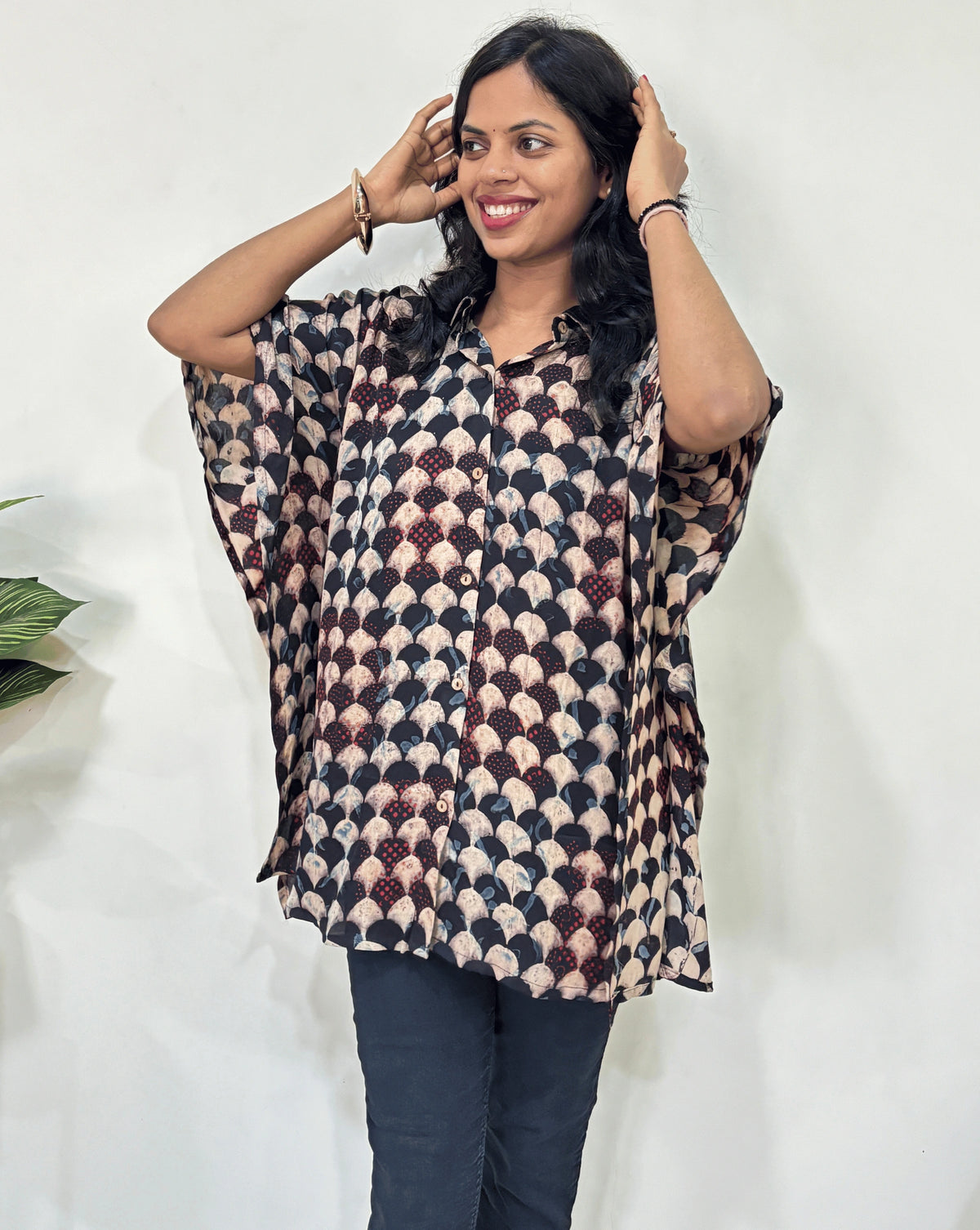 Front view of Beige Women's Ajrakh pure Mashru Silk Short Kaftan Shirt Featured with Collar neck & Elbow sleeves with a bold abstract of black, and red geometric print, paired with dark pants, standing in front of a white wall.