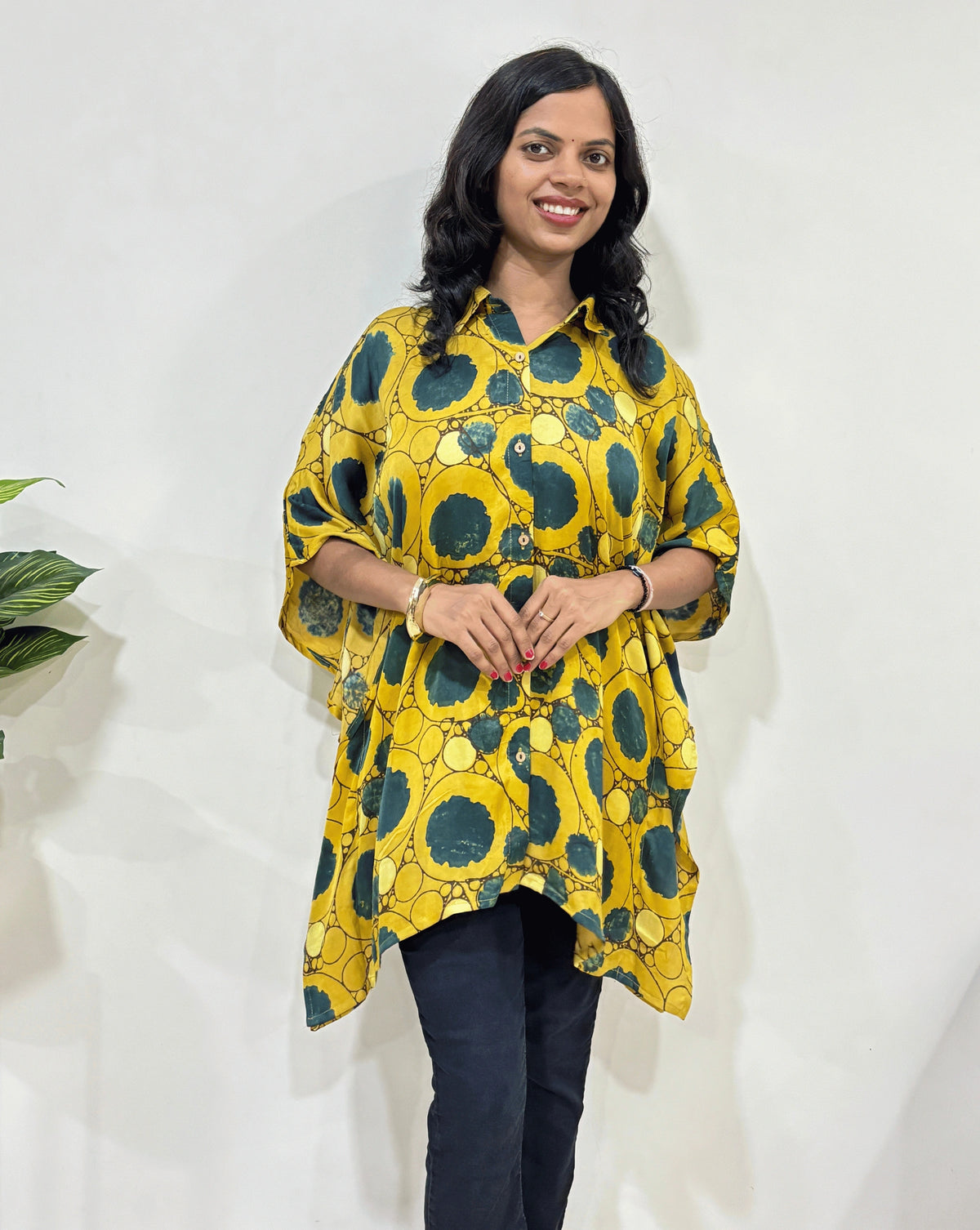 Front view of Yellow Women's Ajrakh pure Mashru Silk Short Kaftan Shirt Featured with Collar neck & Elbow sleeves with a bold abstract of Green , black, and Light yellow, geometric circles print, paired with dark pants, standing in front of a white wall.