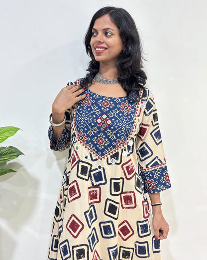 Close-up of Beige Women's Ajrakh Vegetable dyed plant based cotton One-peice Featured three-quarter Sleeves & Round Neck, with beautiful patola print yoke on front, bottom & Sleeves , Geometric print on front & sleeves Hand-mirror work on front chest, pocket & Sleeves cuff, Standing in front of White wall.