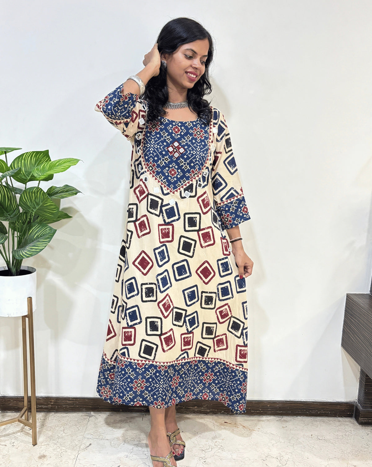 Front View of Beige Women's Ajrakh Vegetable dyed plant based cotton One-peice with beautiful patola print yoke on front, bottom & Sleeves , Geometric print on front & sleeves Hand-mirror work on front chest, pocket & Sleeves cuff, Standing in front of White wall.