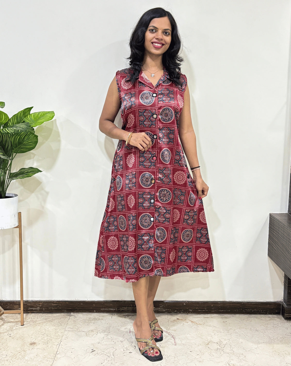Front view of Red Women's Ajrakh pure Mashru Silk Long Kurti Featured with Collar neck & Sleeveless, with a base Red & Abstract White & Blue Geometric Bandhani print, Standing in front of white wall, left side green plant pot.