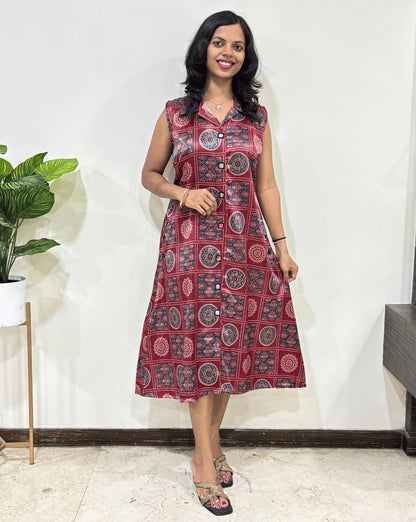 Front view of Red Women's Ajrakh pure Mashru Silk Long Kurti Featured with Collar neck & Sleeveless, with a base Red & Abstract White & Blue Geometric Bandhani print, Standing in front of white wall, left side green plant pot.