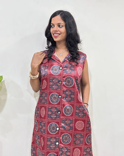 Close-up of Red Women's Ajrakh pure Mashru Silk Long Kurti Featured with Collar neck & Sleeveless, with a base Blue & Abstract White & Red Geometric Bandhani print, A-line pattern, 2 side pockets, Hand-mirror work on front & pockets, Standing in front of white wall, left side green plant pot.
