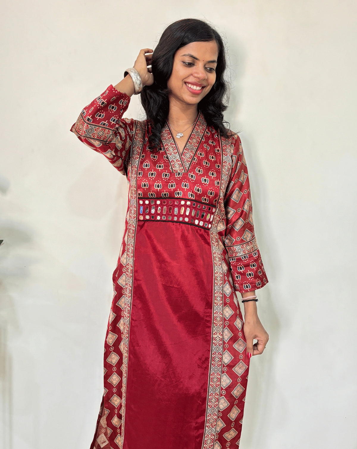 Close-up of Red Woman's Ajrakh Pure Mashru Silk Long-Kurti featured V-shaped neck & Three-Quarter Sleeves with Ajrakh print panel-border on front & Sleeves cuffs, Hand-mirror work on front, patterned dress with a red center , geometric print outside , standing against a white wall