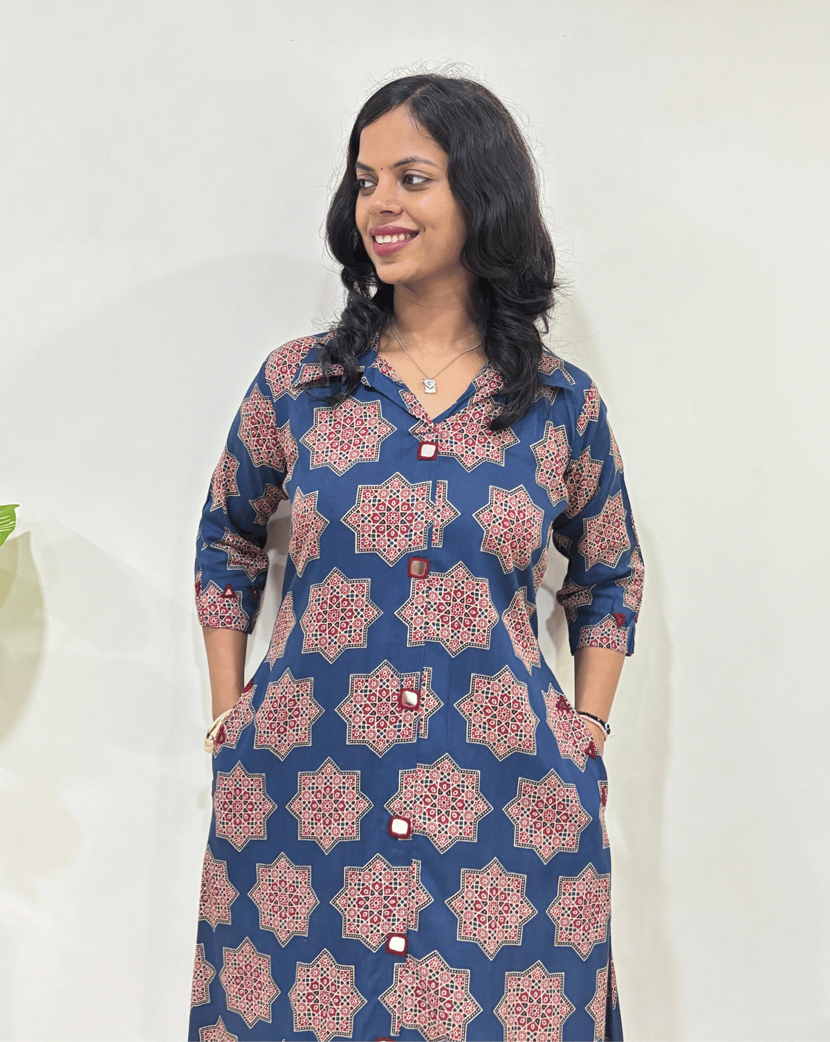 Close-up of Blue women's Ajrakh vegetable dyed plant based cotton Long kurti Featured with three-Quarter Sleeves with a blue base & abstract red Star print, Hand-mirror work on Front & Sleeves cuffs, with 2 side pockets, standing indoors with a white wall and plant in the background