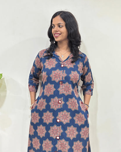 Close-up of Blue women's Ajrakh vegetable dyed plant based cotton Long kurti Featured with three-Quarter Sleeves with a blue base & abstract red Star print, Hand-mirror work on Front & Sleeves cuffs, with 2 side pockets, standing indoors with a white wall and plant in the background