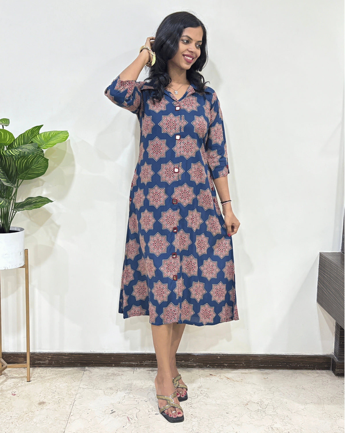 Front View of Blue women's Ajrakh vegetable dyed plant based cotton Long kurti with a blue base & abstract red Star print, Hand-mirror work on Front & Sleeves cuffs, standing indoors with a white wall and plant in the background