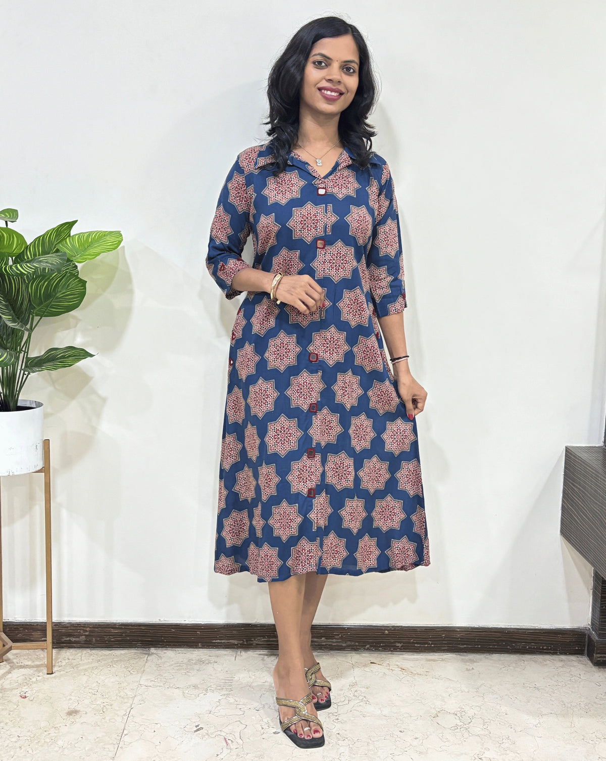 Blue women's Ajrakh vegetable dyed plant based cotton Long kurti with a blue base & abstract red Star print, standing indoors with a white wall and plant in the background